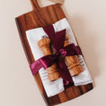 Load image into Gallery viewer, Closing gift with an acacia wood paddle cutting board, olive wood salt and pepper mills, and white striped tea towels wrapped in burgundy satin ribbon