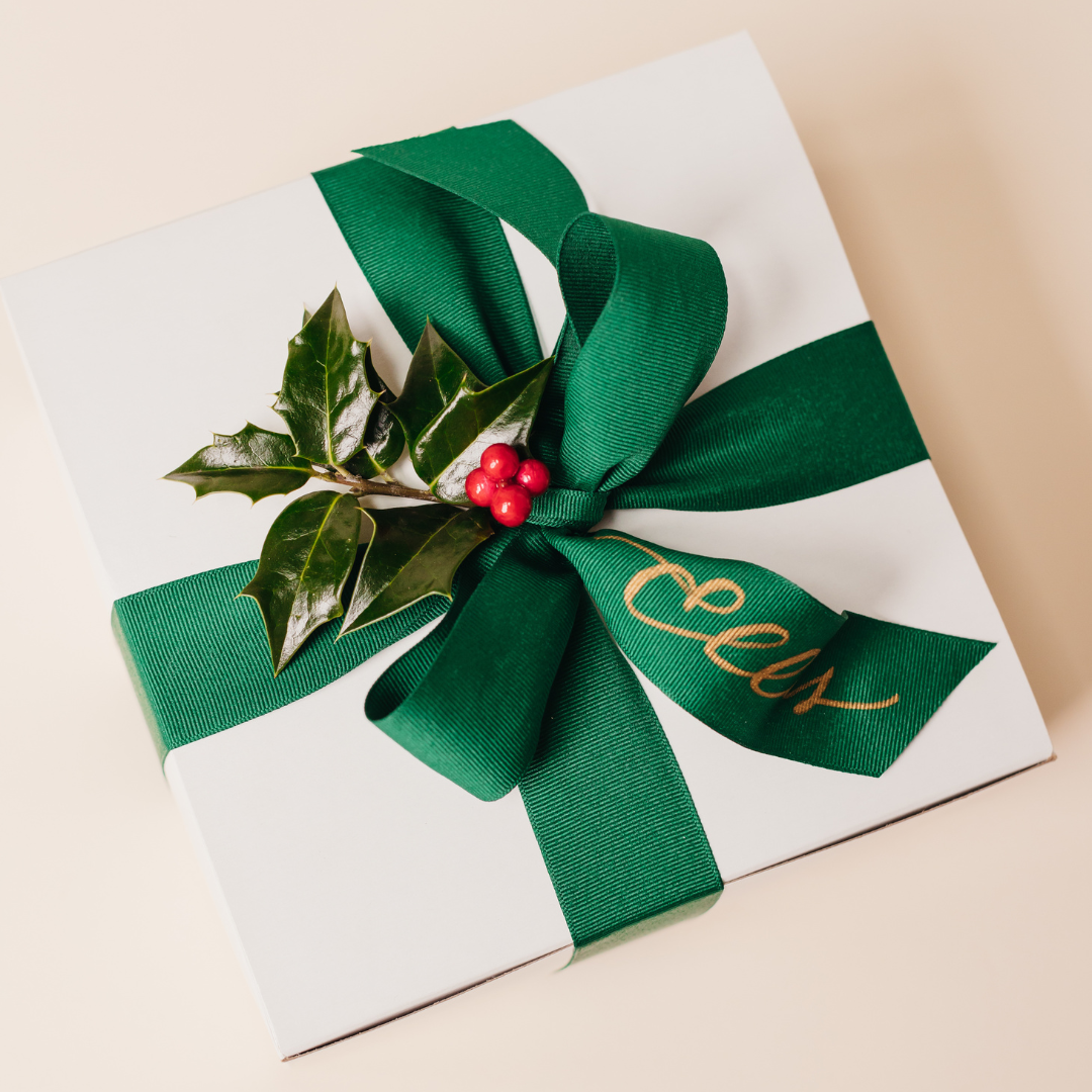 Custom corporate holiday gift box with green ribbon bow, holly accent, and gold-foil name ribbon, an example of Lavender + Pine's corporate gifting program.