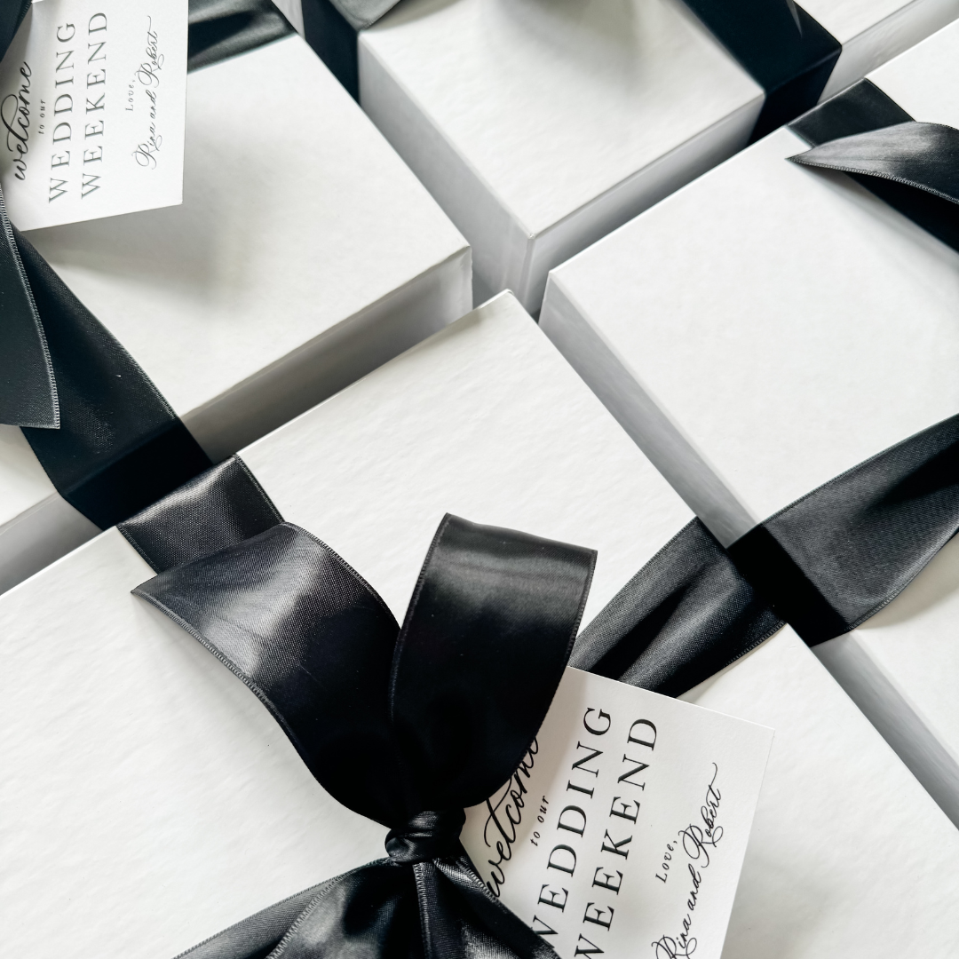 White wedding welcome gift boxes tied with black satin ribbon and personalized gift tags reading "Welcome to the Wedding Weekend