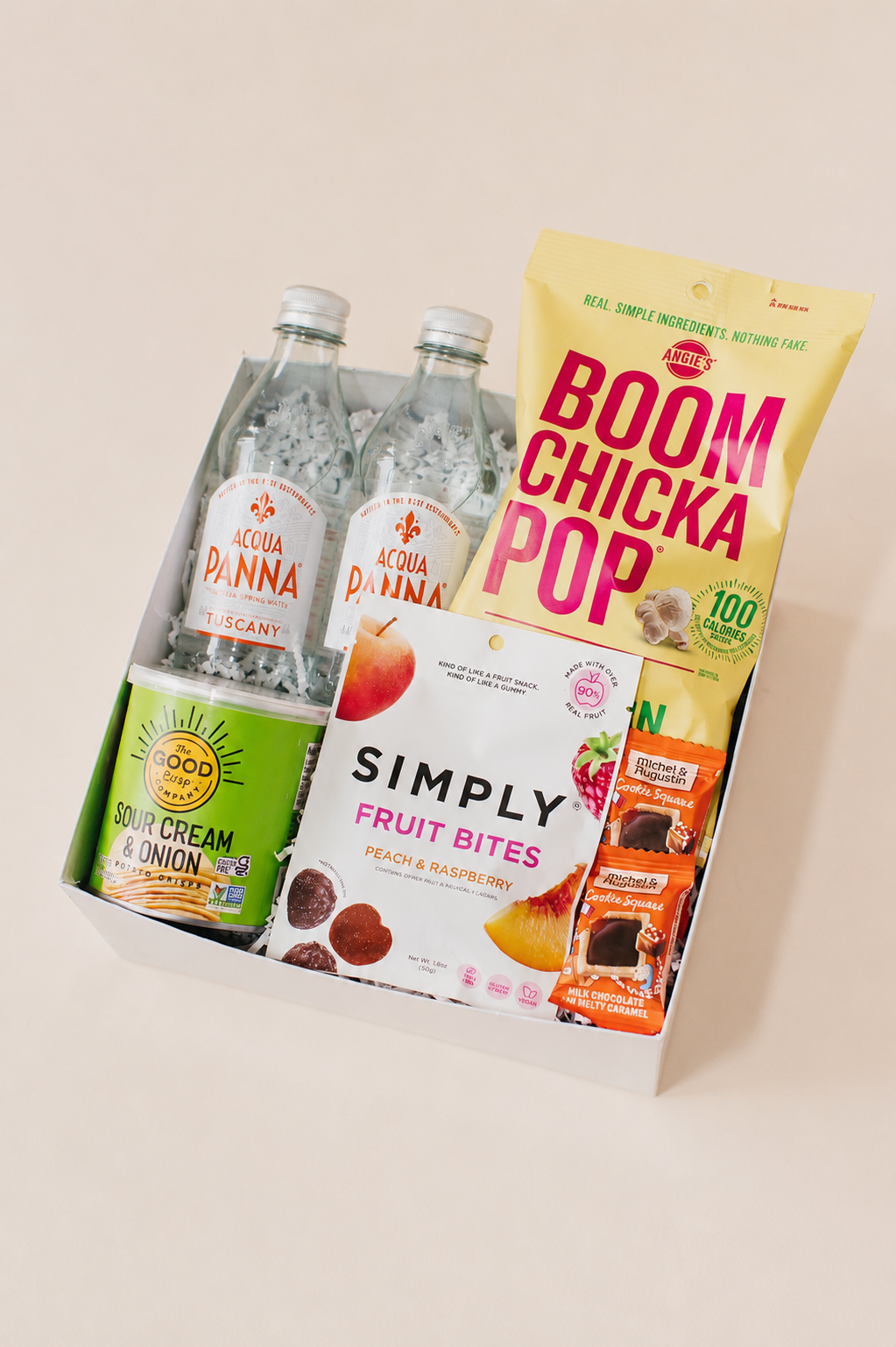 Assorted snacks for summer welcome bags including bottles, a box of fruit bites, and a bag of Boom Chicka Pop on a light background
