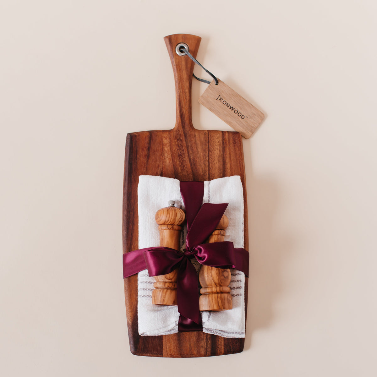 Housewarming gift set with an Ironwood acacia wood paddle cutting board, olive wood salt and pepper mills, and striped linen tea towels tied with burgundy satin ribbon
