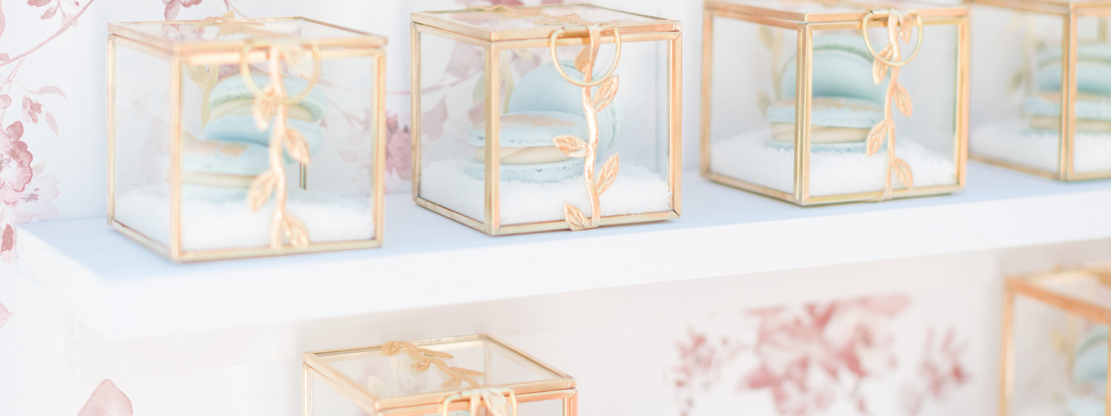 Glass and gold favor boxes displayed on a shelf, each holding pastel macarons with decorative gold leaf detailing
