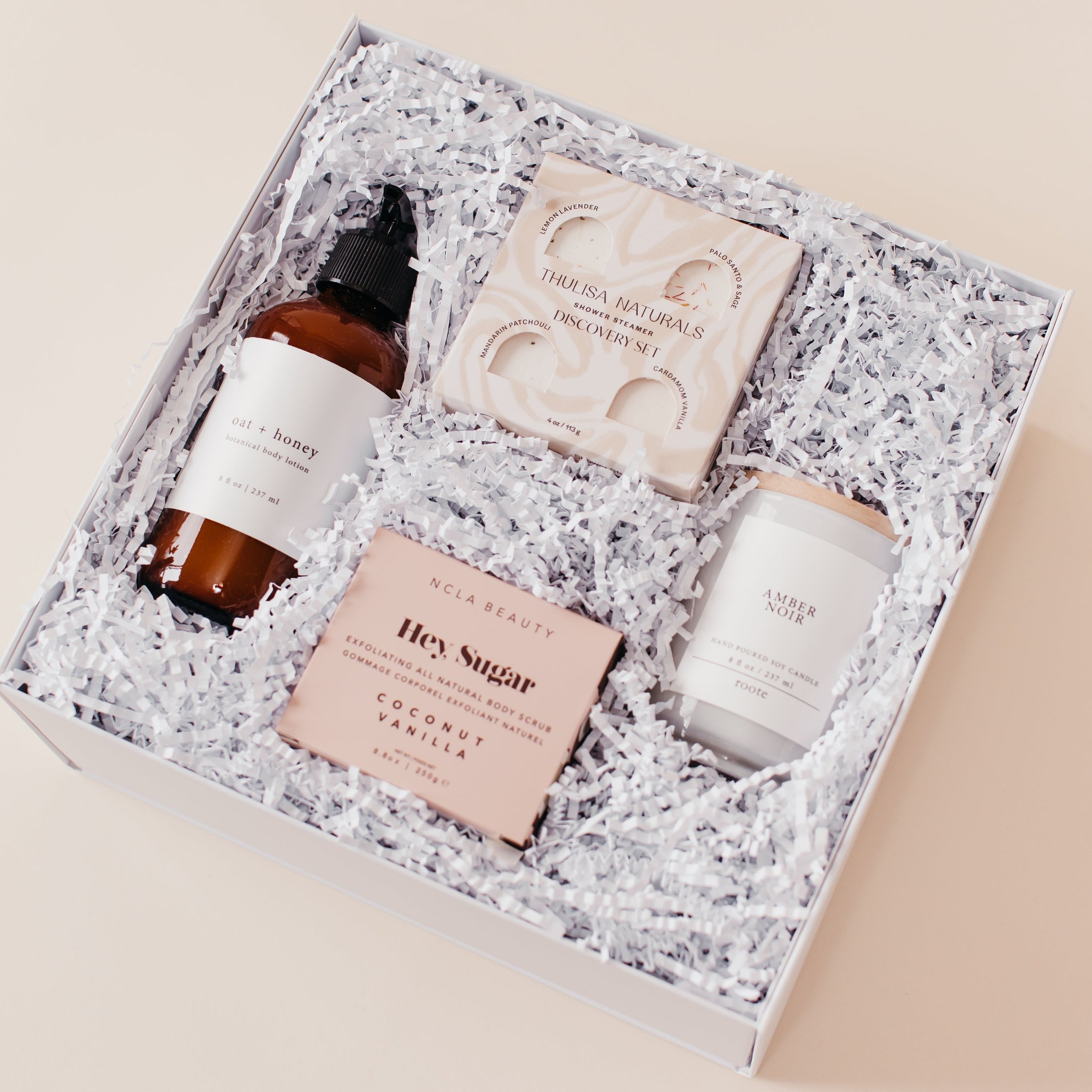 Lavender and Pine Self Care Sunday Gift Box angle view featuring Roote oat and honey body lotion, Thulisa Naturals shower steamer discovery set, NCLA Beauty Hey Sugar coconut vanilla body scrub, and Roote Amber Noir candle in white luxe two piece gift box