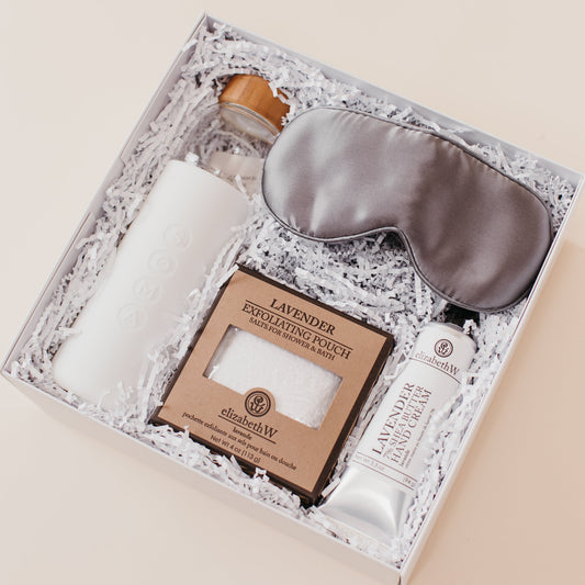 Lavender and Pine Soft Reset Gift Box angle view featuring Soma glass water bottle, Pure Sol silk sleep mask, ElizabethW lavender exfoliating shower pouch, and ElizabethW lavender shea butter hand cream in white luxe two piece gift box