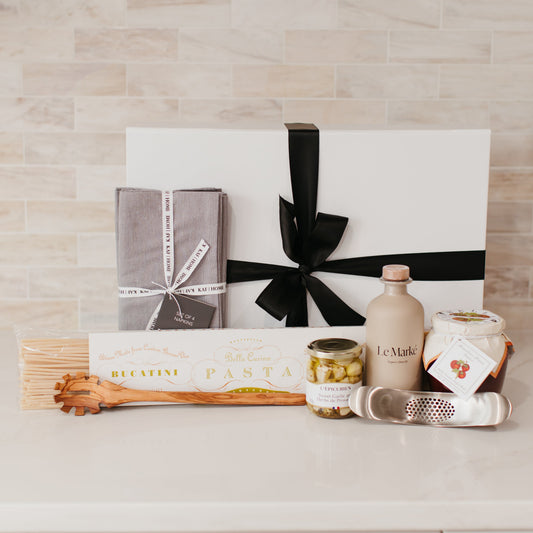 Lavender and Pine Sunday Sauce Gift Box detail view featuring Bella Cucina bucatini pasta, Le Marké olive oil, French Farm L'Epicurien sweet garlic and herbs, Bella Cucina arrabbiata sauce, stainless garlic rocker, Natural OliveWood pasta spoon, and Kaf Home grey linen napkins in white magnetic gift box