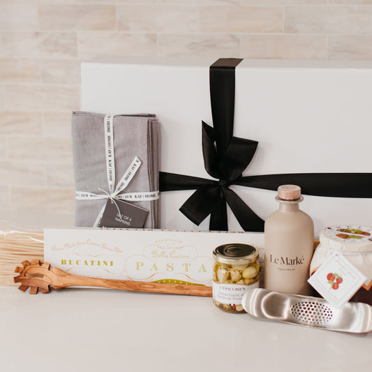 Lavender and Pine Sunday Sauce Gift Box front view featuring Bella Cucina bucatini pasta, Le Marké olive oil, French Farm L'Epicurien sweet garlic and herbs, Bella Cucina arrabbiata sauce, stainless garlic rocker, Natural OliveWood pasta spoon, and Kaf Home grey linen napkins in white magnetic gift box