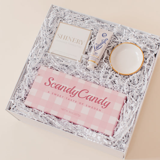 Lavender and Pine Sweet Surprise Gift Box angle view featuring Scandy Candy Swedish candy, Shinery radiance towelettes, Lanolips 101 ointment, and Apricity Ceramics gold rim ring dish in white luxe two piece gift box
